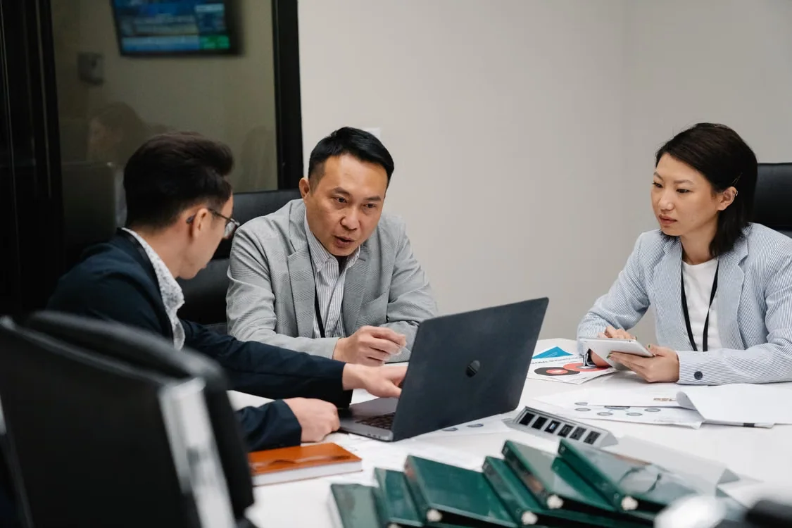 Three business professionals reviewing export documents around a laptop during a logistics meeting